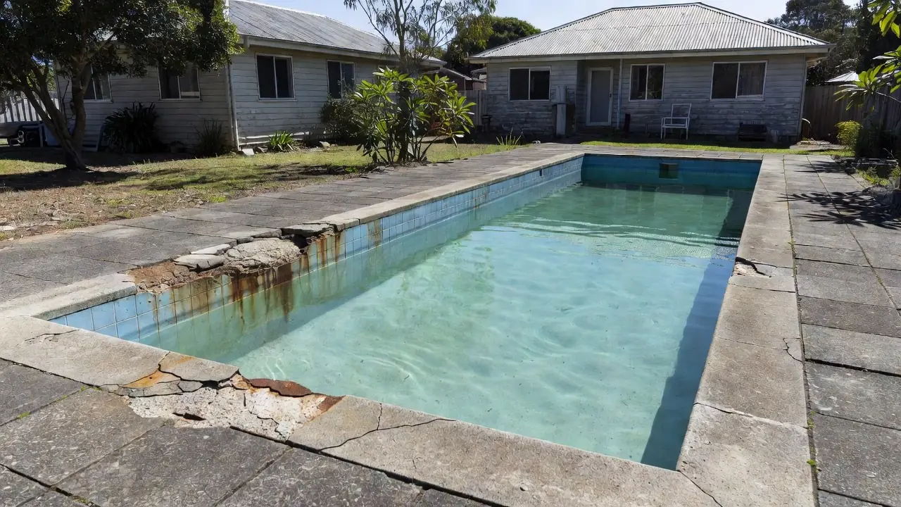 A spalled concrete pool coping beam with rust bleeding into the tiles.