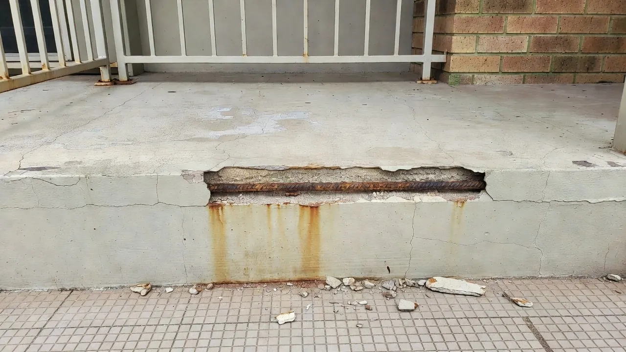 A balcony slab edge where a length of concrete cover has broken off, showing a rusted bar inside, with rust stains below and fragments on the tiles.